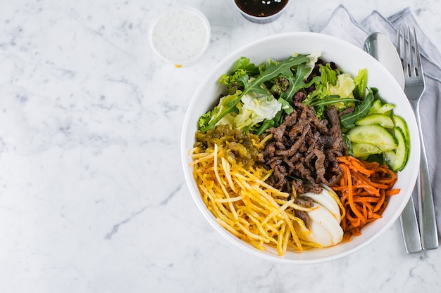 Tazón de almuerzo asiático con carne de res. fideos, mezclar hojas verdes y vegetales en la mesa de mármol. Vista superior, espacio de copia