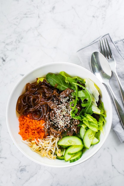 Tazón de almuerzo asiático con carne de res. fideos, mezclar hojas verdes y vegetales en la mesa de mármol. Vista superior, espacio de copia