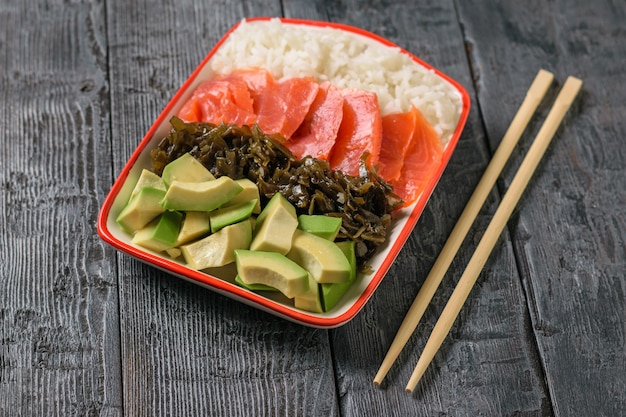 Tazón con arroz, algas, rodajas de aguacate y pescado en una mesa negra con palos de madera.