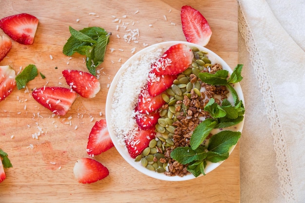 Tazón de batido de bayas de desayuno cubierto con muesli de coco de semillas de calabaza de fresa y menta fresca Tazón de batido de plátano de fresa desayuno saludable o merienda vajilla de madera