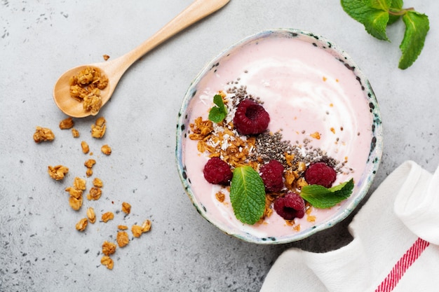 Tazón de batido de plátano y frambuesa rosa con granola, semillas de chía, coco y menta sobre hormigón gris claro.