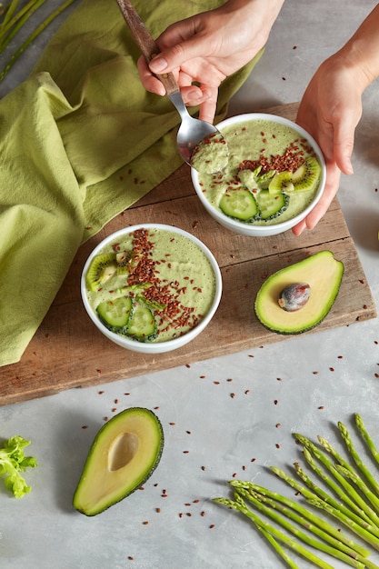 Tazón de batido de superalimento verde con kiwi, espárragos, pepino, aguacate y semillas flexibles en las manos del wooman sobre tabla de madera con servilleta verde sobre fondo de piedra. Endecha plana