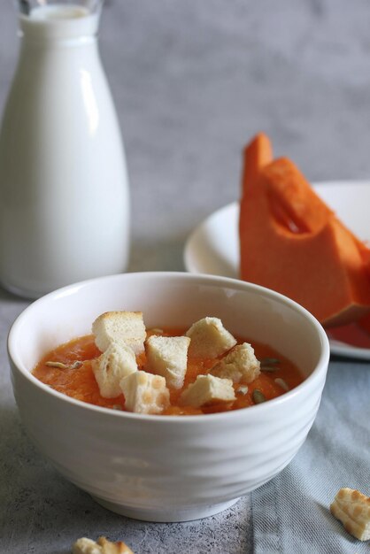 Un tazón blanco con sopa de crema de calabaza, picatostes y semillas, una botella de leche y una rodaja de calabaza sobre un fondo claro. Vista lateral. Orientación vertical.