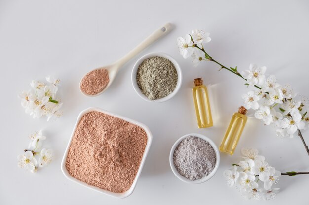 Tazón de cerámica con polvo de arcilla roja, ingredientes para mascarilla facial y corporal casera o exfoliante y una ramita fresca de cereza en flor sobre una superficie blanca. Concepto de spa y cuidado corporal.