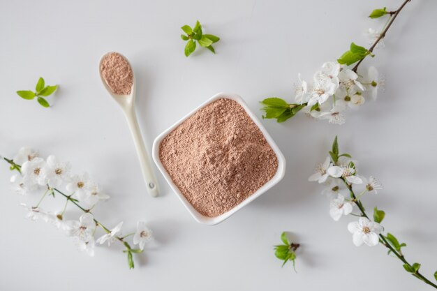 Tazón de cerámica con polvo de arcilla roja, para mascarilla facial y corporal casera o exfoliante y una ramita fresca de cereza en flor sobre una superficie blanca. Concepto de spa y cuidado corporal.