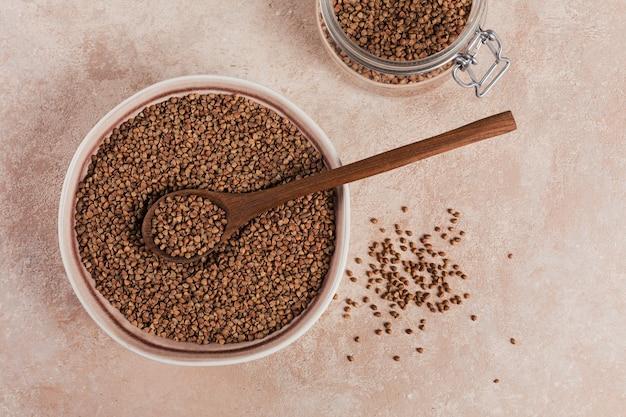 Tazón de cereales secos de trigo sarraceno crudo con cuchara de madera sobre un fondo beige claro