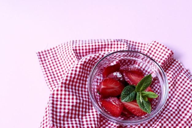 Tazón de deliciosas fresas en rodajas con detalle de hierba de menta y mantel a cuadros rojo y blanco y fondo rosa con espacio de copia