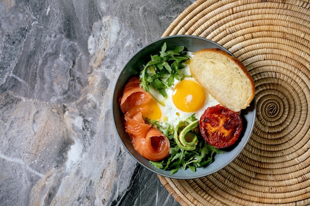 Tazón de desayuno saludable con huevos fritos, salmón, aguacate, tomate a la parrilla y ensalada que se sirve con pan en una servilleta de paja. Endecha plana