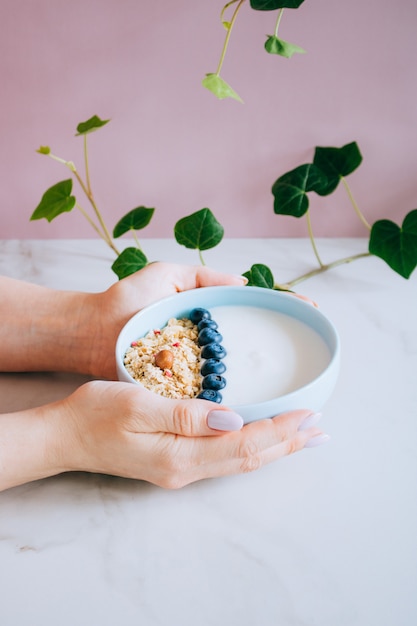 Tazón de desayuno saludable con muesli y yogurt en manos de mujer sobre fondo rosa y mármol