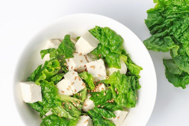 Tazón con ensalada verde y rodajas de tofu, en la pared blanca, vista superior