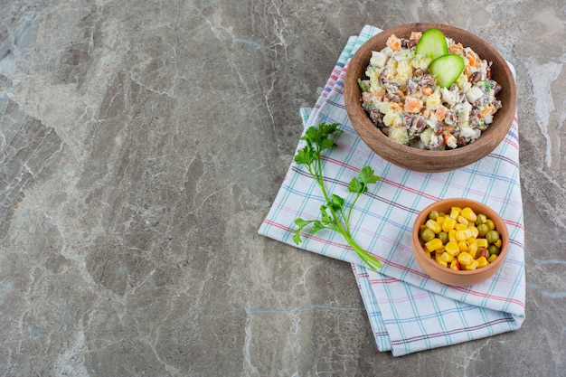 Un tazón de ensalada de verduras junto a una ensalada de maíz en un tazón sobre un paño de cocina, sobre el fondo de mármol.