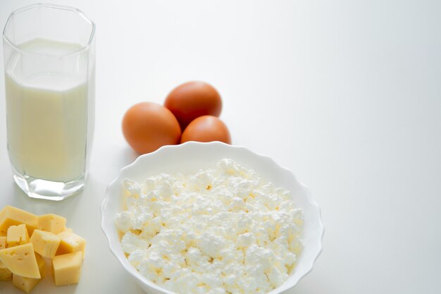Un tazón de fuente blanco con huevos de requesón queso cortado en cubitos y un vaso de leche de pie sobre un fondo blanco ...