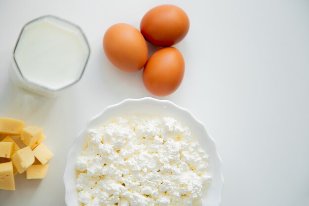 Un tazón de fuente blanco con huevos de requesón queso cortado en cubitos y un vaso de leche de pie sobre un fondo blanco ...