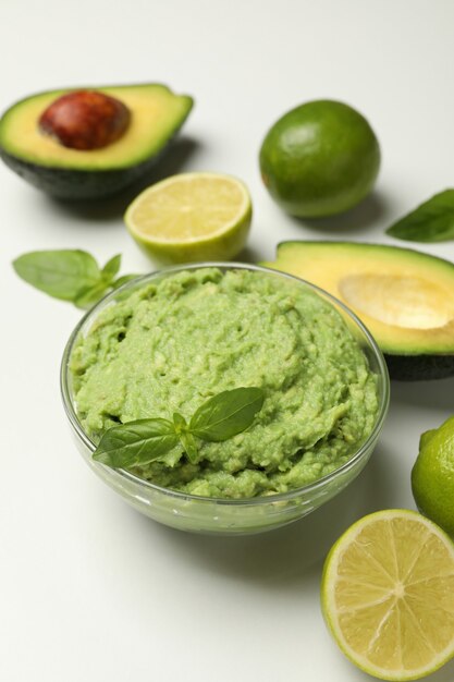 Tazón de fuente de guacamole, aguacate, lima y albahaca sobre fondo blanco.