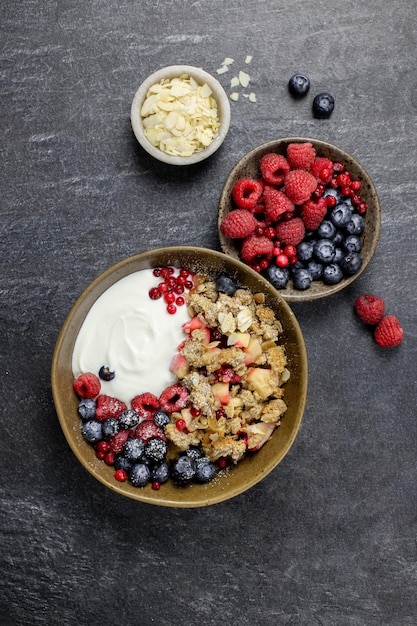 Tazón de granola casera con yogur y bayas frescas sobre fondo de hormigón de piedra oscura. Crumble de arándano y manzana, crujiente con copos de almendra y avena. Vista superior.