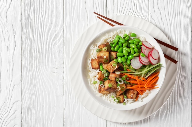 Tazón de tofu vegano con arroz basmati, frijoles edamame, rábano, pepino en rodajas finas y zanahorias en un tazón blanco con palillos, vista horizontal desde arriba, primer plano, espacio libre