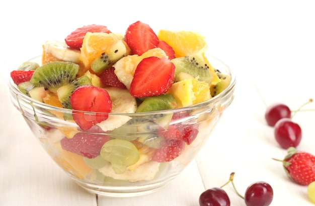 Tazón de vidrio con ensalada de frutas frescas y bayas en la mesa de madera blanca