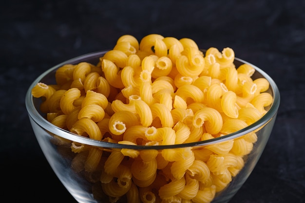 Tazón de vidrio con pasta rizada de trigo dorado sin cocer cavatappi sobre fondo negro oscuro con textura, ángulo de visión macro