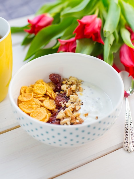 Tazón de yogur delicioso con copos de maíz, nueces y mermelada en una mesa de madera blanca. Concepto de nutrición saludable y orgánica. Tulipanes con taza de té y desayuno.