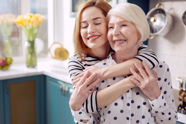Te amo. Encantadora mujer mayor dando a su anciana madre un abrazo por la espalda, cerrando los ojos con placer y sonriendo felizmente mientras está de pie en la cocina