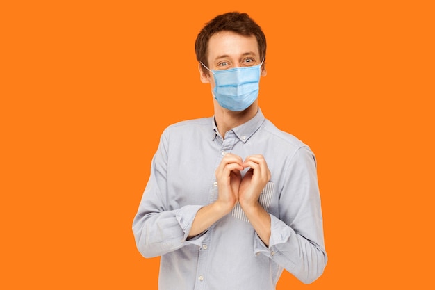 Te amo. Retrato de hombre joven trabajador feliz alegre con máscara médica quirúrgica de pie con gesto de amor de corazón y mirando a cámara con una sonrisa. tiro de estudio de interior aislado sobre fondo naranja.