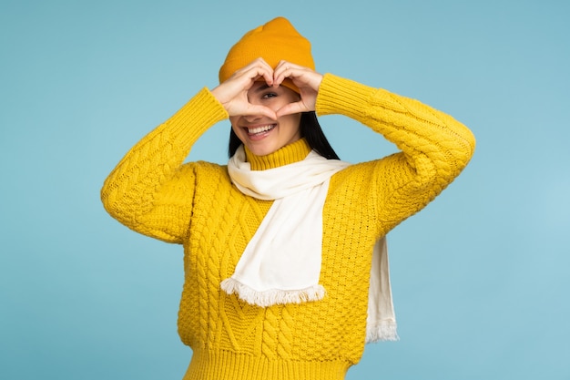 Te amo. Retrato de mujer joven romántica atractiva con cabello castaño de pie y haciendo corazón con las manos, sonriendo juguetonamente. Tiro de estudio interior aislado sobre fondo azul.