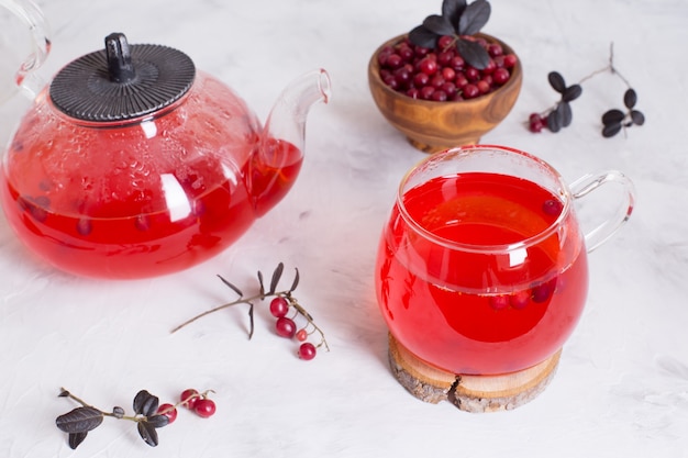 Té de arándano caliente en una taza de vidrio y una tetera sobre un fondo claro.