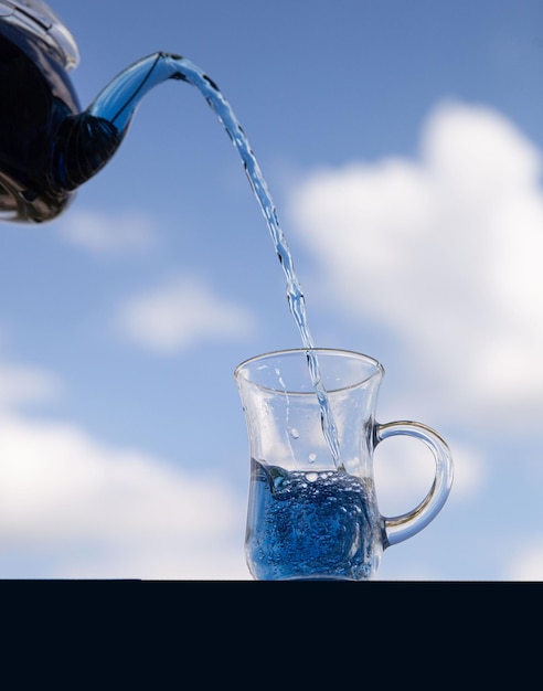 El té azul floral se vierte de una tetera en un vaso de vidrio contra el fondo del cielo con nubes