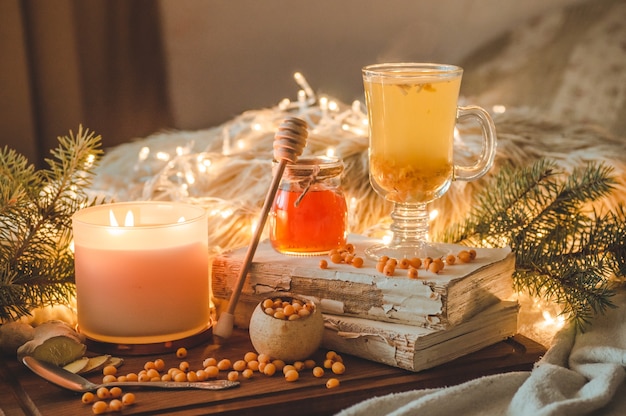 Té con bayas de espino amarillo y jengibre sobre libros antiguos, miel, velas y ramas de coníferas. El ambiente de confort en casa. Hogar cálido y acogedor.