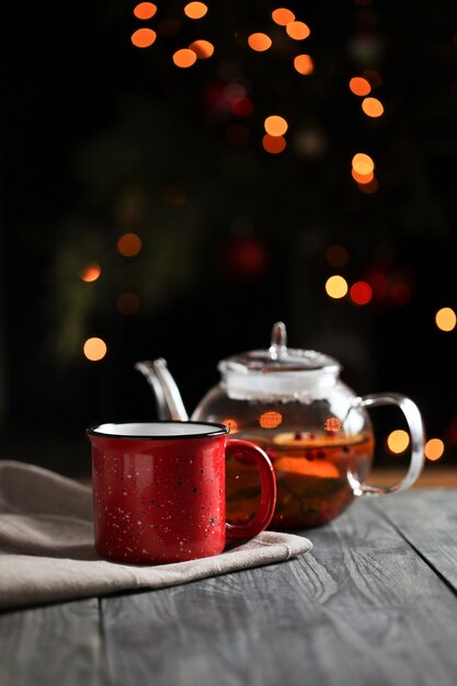 Té caliente en una tetera de cristal junto a una taza roja decorada con una rodaja de naranja por la mañana sobre un fondo de luces. Rodajas de naranja sobre un fondo oscuro.