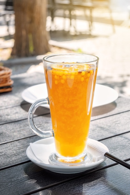 Té de espino amarillo naranja en una taza de vidrio a la luz del sol en un restaurante de la calle. Bebida amarilla sobre la mesa en un café del parque.