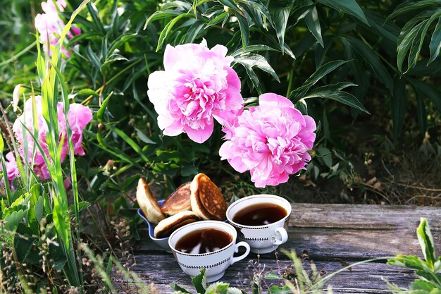 Té en estilo campestre en el jardín de verano en el pueblo Dos tazas de té negro y panqueques en tablas de madera y flores de peonía florecientes a la luz del sol