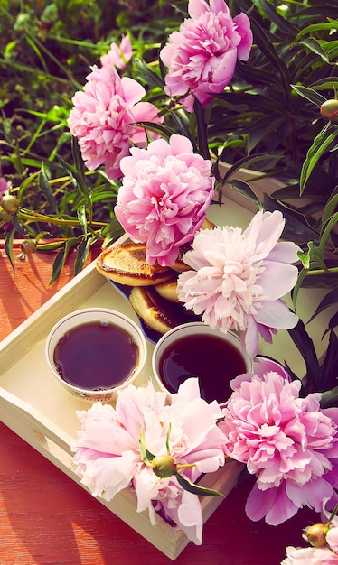 Té de estilo rústico en el jardín de verano del pueblo. Dos tazas de té negro en una bandeja de madera y flores de peonía florecientes a la luz del sol.