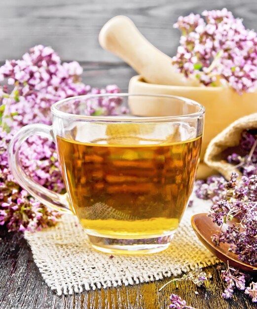 Té de hierbas de orégano en una taza de vidrio sobre arpillera, flores frescas en mortero y sobre la mesa, flores secas de mejorana en una bolsa y cuchara sobre fondo de tablero de madera