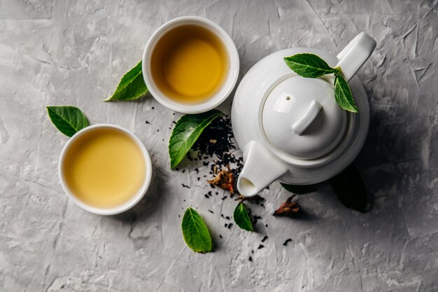 Té de hierbas, tazas y tetera con hojas sobre fondo de hormigón gris.