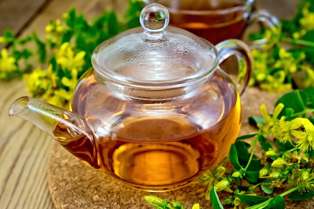 Té de hierbas en una tetera de vidrio en el soporte, taza, flores frescas tutsan en el fondo de tablas de madera