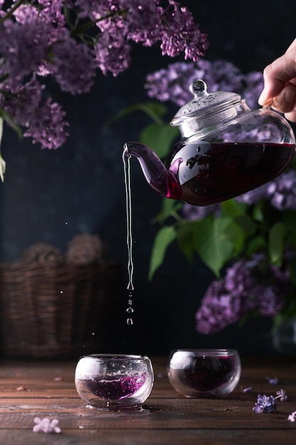 El té morado se vierte de una tetera de vidrio en tazas. Las flores lilas están por todas partes.