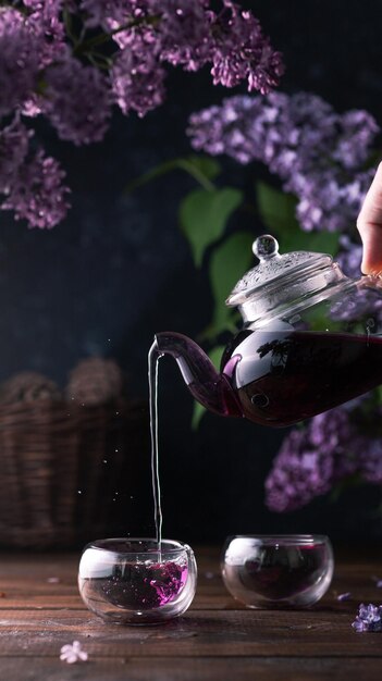 El té morado se vierte de una tetera de vidrio en tazas. Las flores lilas están por todas partes.