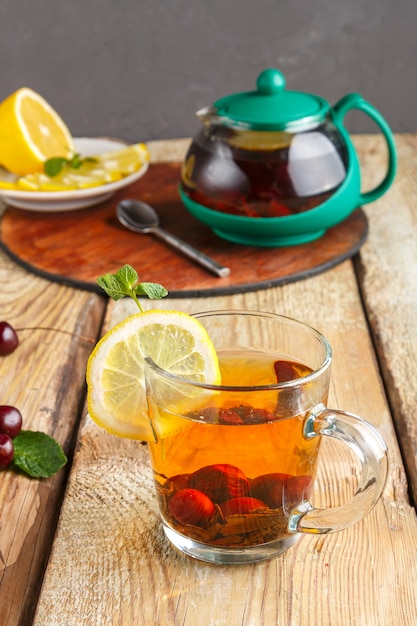 Foto té negro en una taza de vidrio con cerezas de menta y limón sobre una mesa de madera y una tetera y limón en un plato y hojas de menta.