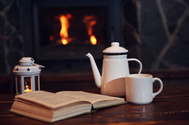 Té o café caliente en taza, libro y velas en la mesa de madera vintage. Chimenea como fondo