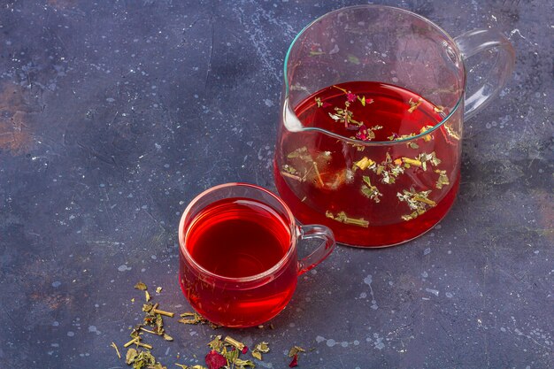 Té rojo (rooibos, hibisco, karkade) en vaso de vidrio y tetera entre hojas de té seco y pétalos sobre un fondo oscuro. Té de hierbas, vitaminas y desintoxicación para el resfriado y la gripe. Cerrar, copiar espacio para texto