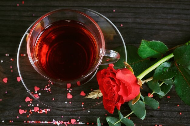 Té en taza transparente, caramelos de colores y una rosa roja sobre la oscuridad.