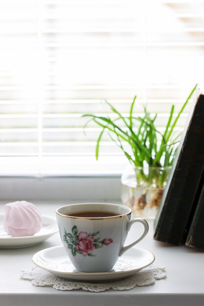 Té en una taza vintage, malvaviscos en un platillo vintage y libros antiguos sobre un fondo claro.