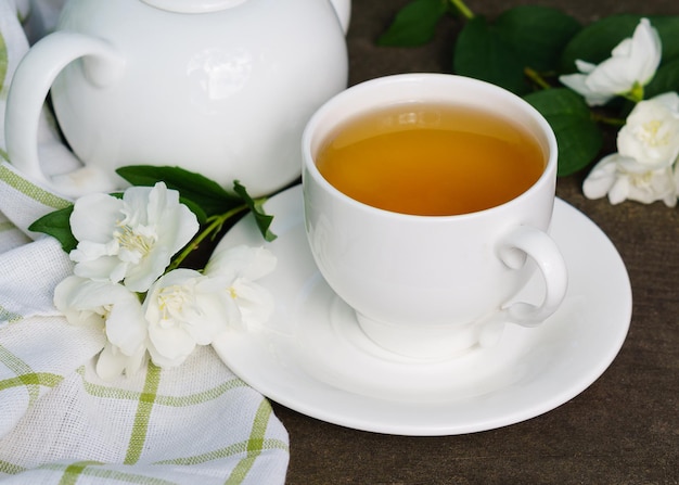 Té verde caliente en una tetera y una taza con una rama de flores de jazmín en flor y una toalla blanca sobre fondo de madera marrón rústico áspero. Concepto de vida lenta saludable de la naturaleza.