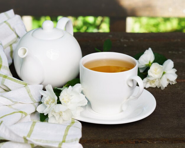 Té verde caliente en una tetera y una taza con una rama de flores de jazmín en flor y una toalla blanca sobre fondo de madera marrón rústico áspero. Concepto de vida lenta saludable de la naturaleza.