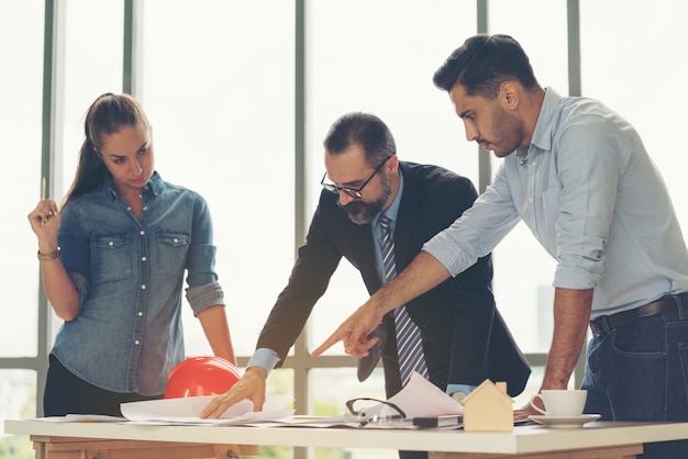 Team multiethnischer Architekten, die im Besprechungsraum an Bauplänen arbeiten. Ingenieure diskutieren über Projekt im Büro. Reifer Geschäftsmann und Frau, die um Tisch stehen, der an Blaupause arbeitet.