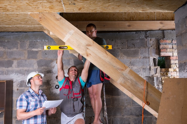 Team von Bauarbeitern, die einen hölzernen Treppenrahmen im unfertigen Keller des neuen Hauses bauen, die Ebenen auf Genauigkeit und Qualitätskontrolle überprüfen