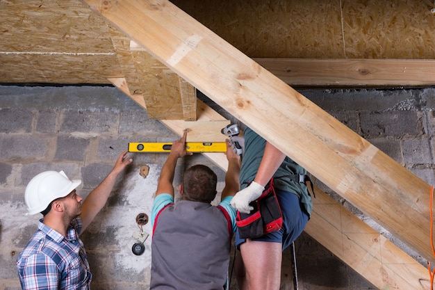 Team von Bauarbeitern, die einen hölzernen Treppenrahmen im unvollendeten Keller des neuen Hauses bauen, die Ebenen auf Genauigkeit und Qualitätskontrolle überprüfen