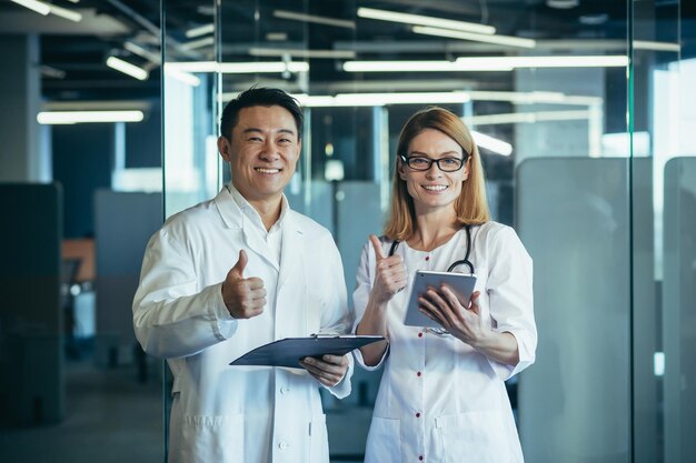 Team von zwei ärzten asiatischer mann und frau, die in die kamera schauen und lächelnd die daumen hochhalten und in einer modernen klinik arbeiten