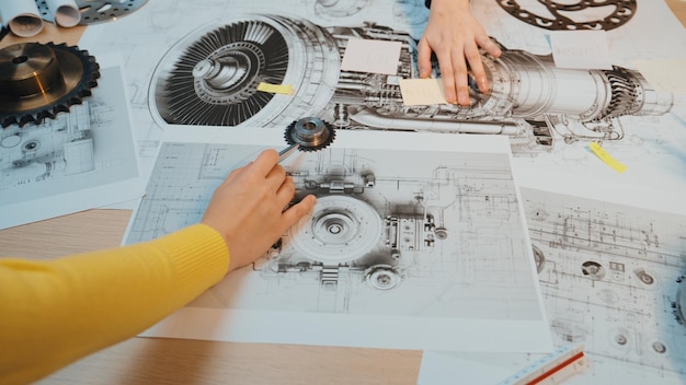 Technischer Zeichnungs- und Konstruktionsprozess mit Händen, die über Schemata mechanischer Teile und Komponenten auf einer Holztischoberfläche interagieren SACTR